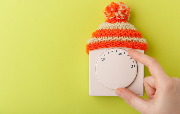 photo of winter hat atop a thermostat that's being turned up by a person's hand