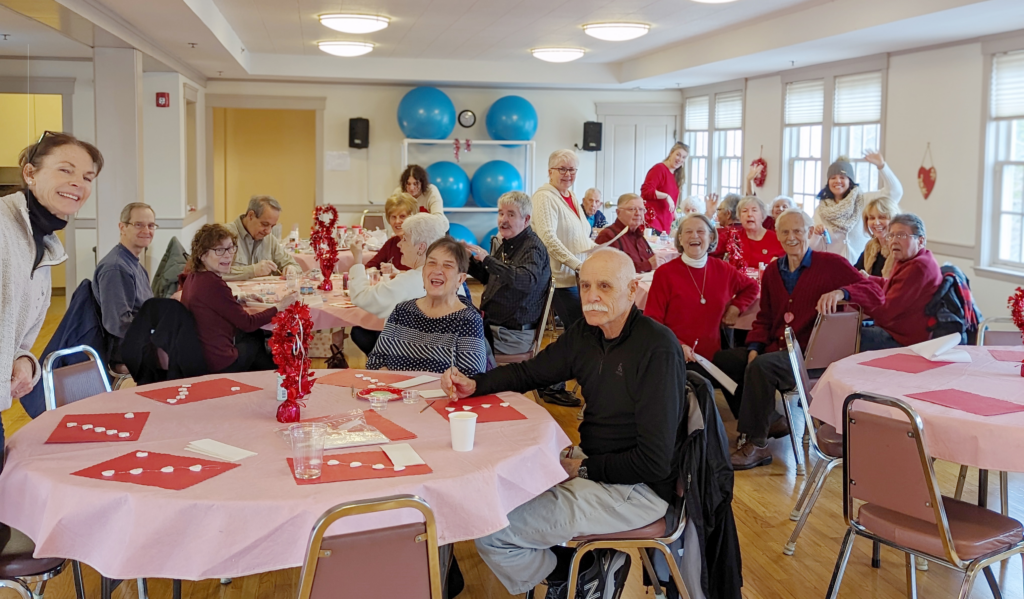 large group of people at various tables, waving to camera or smiling