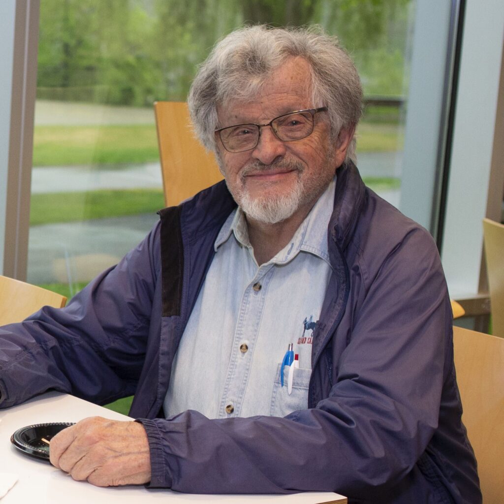 Man with grey hair, grey beard and glasses seen from the shoulders up at an indoor event