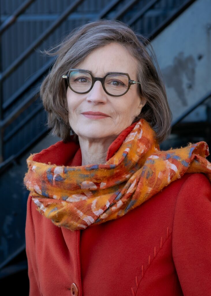 Portrait of a woman with short grey-brown hair, wearing chunky eyeglasses, a red jacket and multicolored decorative scarf, looking at the camera and smiling slightly