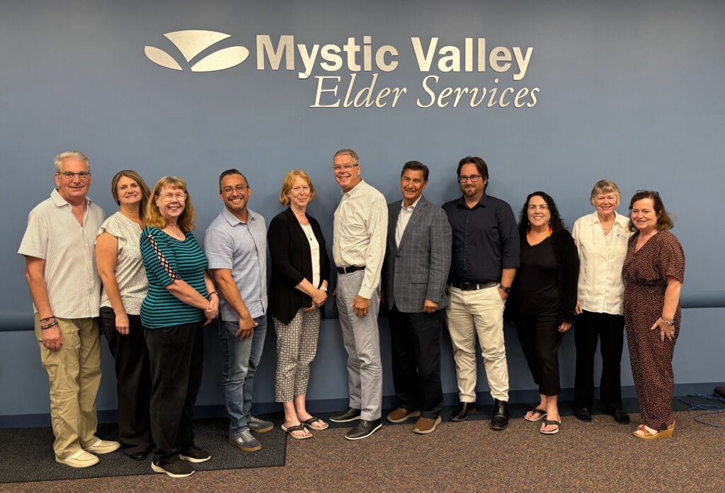 11 men and women stand smiling at the camera. Above them, in silver lettering against a blue wall, is the Mystic Valley Elder Services logo.