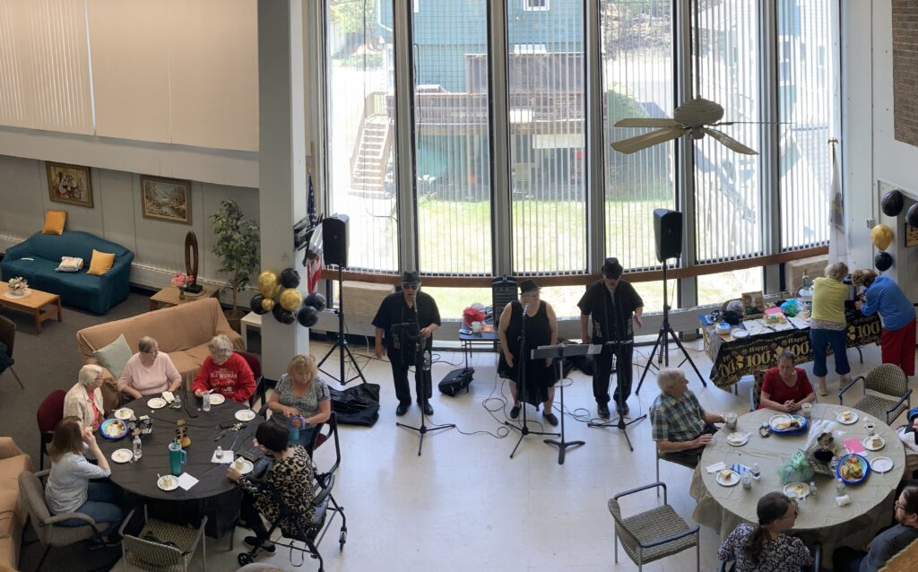 Common room with high windows and ceiling. People are tables and a small band playing in the center.