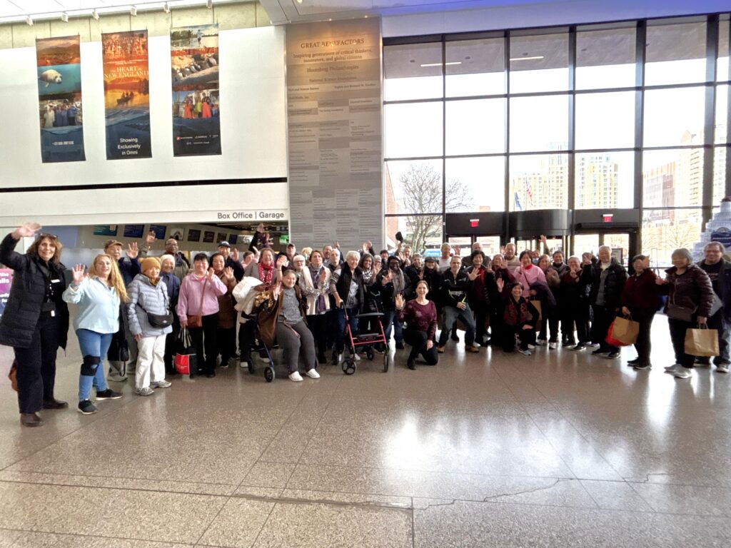 Dozens of older adults and adults with disabilities, along with Mystic Valley Elder Services staff, smile and wave at the camera while in the lobby of the Museum of Science.